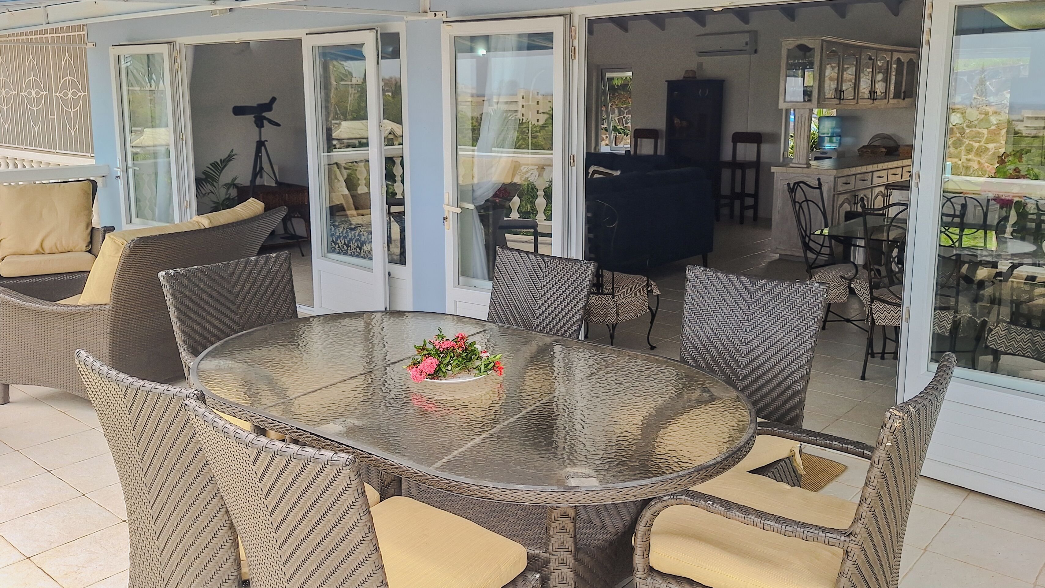 Un patio cubierto con una mesa de vidrio rodeada de sillas de mimbre, un sofá azul oscuro y una zona de comedor con sillas y un armario.
