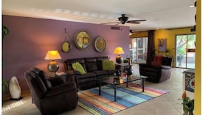 Un salon comprenant un canapé, deux fauteuils, une table basse et des lampes. Les murs sont décorés d’assiettes et le plafond est équipé d’un ventilateur.