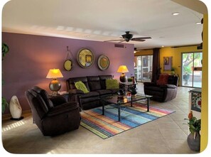 Ein Wohnzimmer mit einer Couch, zwei Sesseln, einem Couchtisch und Lampen. An der Wand befinden sich dekorative Teller und ein Deckenventilator.