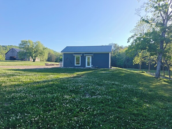 A small, single-story house with a dark exterior and a front door, surrounded by a grassy area with white flowers and trees. - Creek Cabin (Maynardville)