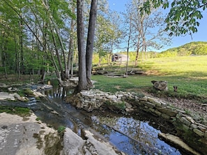 Un paysage naturel avec un ruisseau, des arbres et un petit bâtiment en arrière-plan.