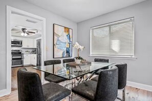 A modern dining area with a glass table, black chairs, a kitchen in the background, and a large window with blinds.