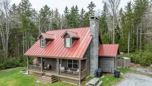 A cabin with a red metal roof, a stone chimney, a porch with seating, and a small shed in a forested area. - Mountain View Retreat (Ludlow)
