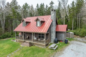 Eine Holzhütte mit rotem Metalldach, einem Steinkamin, einer Veranda mit Sitzgelegenheit und einem kleinen Schuppen in bewaldeter Landschaft.
