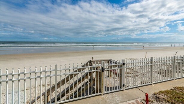 A beach with a white fence, a staircase leading to a wooden structure, and a clear sky. - Delightful, Cozy Modern Apartment By The Ocean @Coastal Waters (New Smyrna Beach)