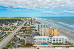 An aerial view of a coastal town with a beachfront hotel, roads, and surrounding greenery. - Delightful, Cozy Modern Apartment By The Ocean @Coastal Waters (New Smyrna Beach)