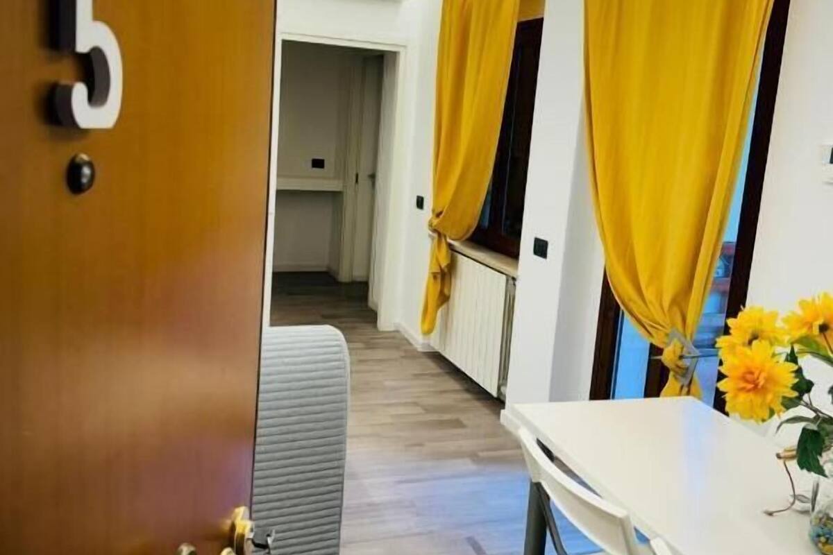 Una camera d'albergo con una porta di legno con il numero '5', un divano grigio, un tavolo bianco e tende gialle.