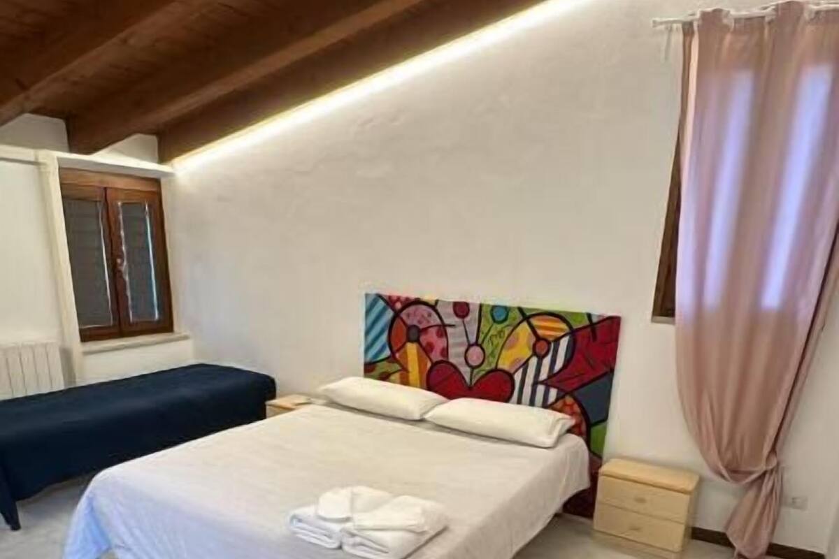 Una camera da letto con un letto, una panca, un quadro colorato, una finestra e una tenda.