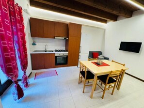A compact kitchen with wooden cabinets, a dining table with chairs, a TV mounted on the wall, and a red curtain. - Fondaco Medievale (Verona)