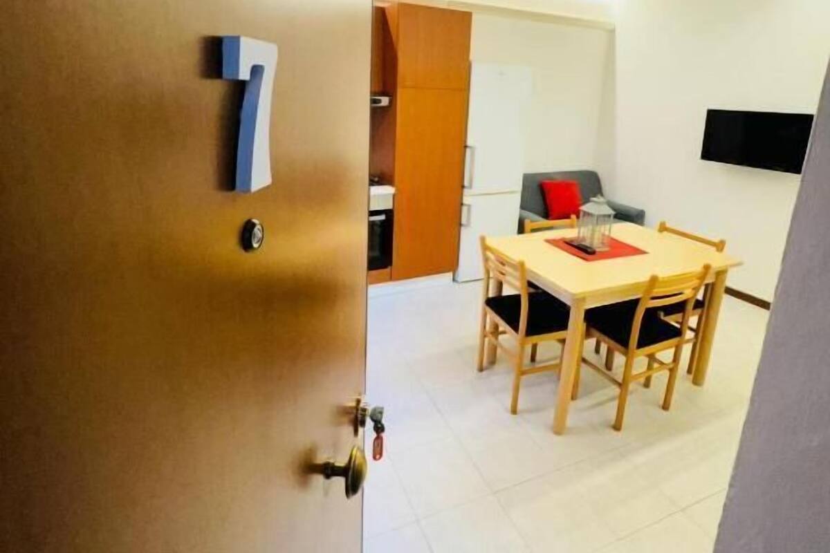 Una stanza con una porta di legno, il numero '7' sulla parete, un tavolo da pranzo con sedie, un televisore montato a parete e un soffitto bianco.