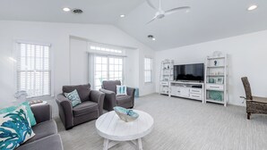 Ein geräumiges Wohnzimmer mit einem weißen runden Couchtisch, grauen Sofas, einem Fernseher und einem Deckenventilator.