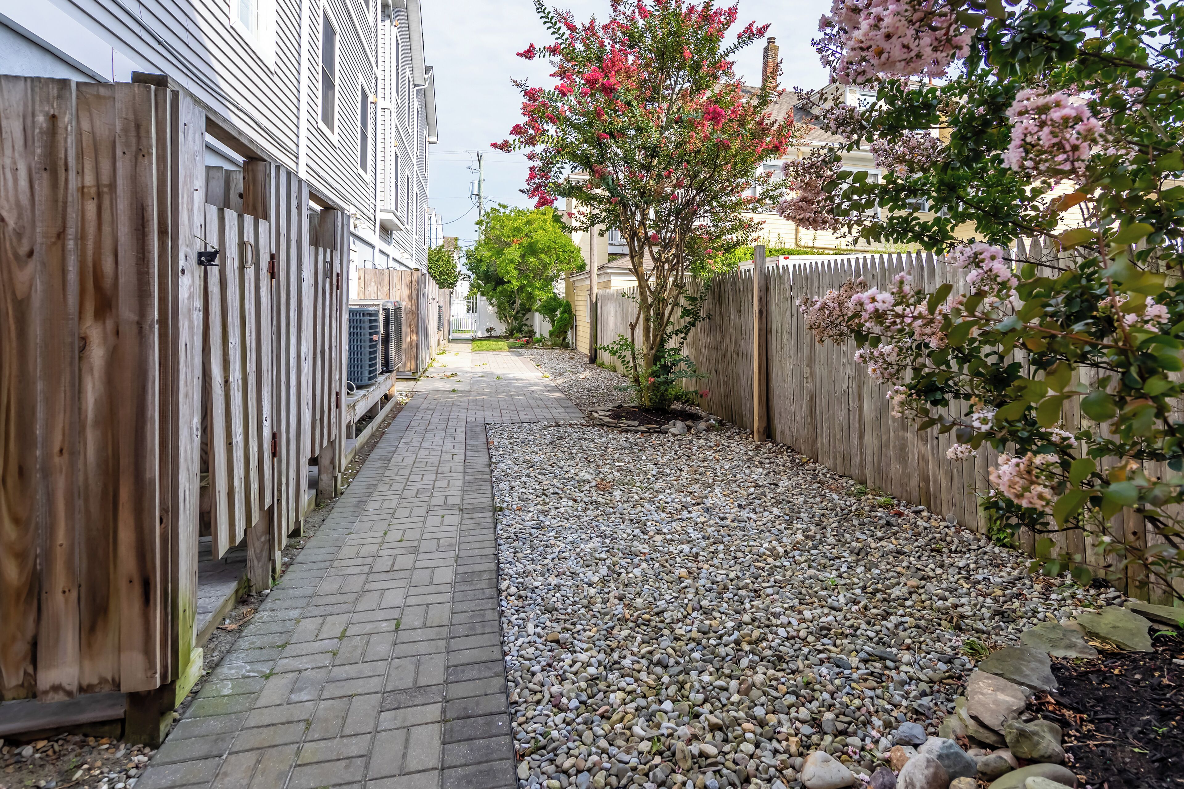 A narrow pathway with a wooden fence on one side and a gravel area on the other, leading to a building with a sloped roof.