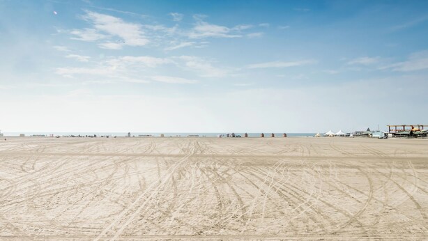 Ein weiter Sandstrand mit Spuren von Reifen, Zelten und einem entfernten Blick auf den Ozean.
