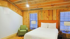 Una habitación con paneles de madera, una cama, una silla y dos ventanas con persianas.