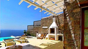 Eine Terrasse mit Steinmauer, einem Holztisch, einem Grill und Blick auf den Ozean.