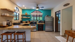 Uma cozinha com ilha central, banquetas, geladeira e ventilador de teto.