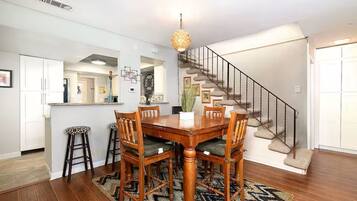 A dining area with a wooden table and chairs, a hanging light fixture, a staircase, and a kitchen area in the background.