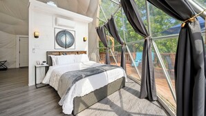 Un dormitorio con cama, mesita de noche, ventilador de pared y un amplio ventanal con cortinas.