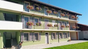 Um edifício de vários andares com varandas decoradas com floreiras, uma escadaria visível e uma área de entrada pavimentada.
