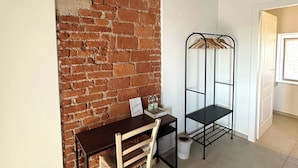 Um quarto com parede de tijolos, uma escrivaninha de madeira com uma cadeira, uma estante de metal e uma porta branca.