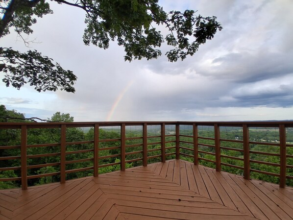 Una terraza de madera con barandilla metálica, con vistas a un paisaje verde exuberante y un arcoíris.