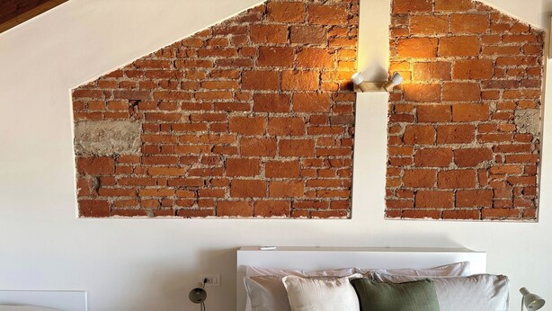Quarto com parede de tijolos, cama com roupa de cama listrada, mesa de cabeceira com abajur e criado-mudo com abajur.