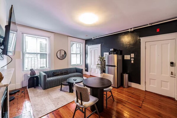 A compact living space with a sofa, dining table, chairs, a small rug, a refrigerator, and a TV.