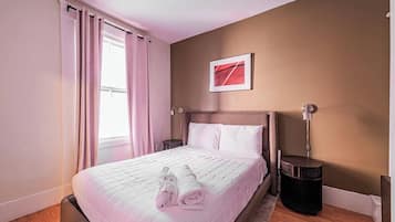 A hotel room with a bed, a bedside table, a lamp, a picture on the wall, and a window with curtains.