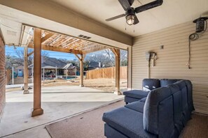 Ein überdachter Patio-Bereich mit Deckenventilator, einem dunkelgrauen Sofa und Blick auf einen Garten mit Holzzaun und Schwimmbecken.