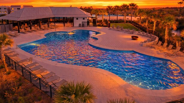 Ein Pool mit geschwungener Form, umgeben von Liegestühlen und einem überdachten Bereich, mit Palmen und einem Sonnenuntergang im Hintergrund.