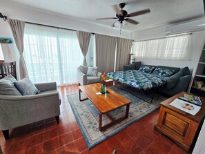 A living room with a sofa, armchair, coffee table, and a bed with a floral bedspread.