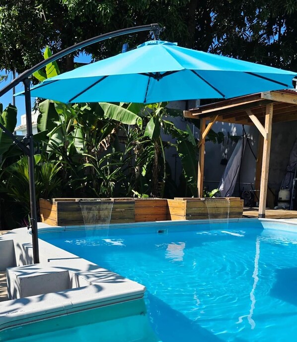 A swimming pool with a blue umbrella, surrounded by lush greenery and a wooden deck.