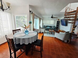 A dining area with a round table, chairs, and a vase with flowers. There is a living room with a sofa and a TV in the background.