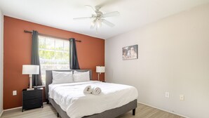 Ein Schlafzimmer mit Bett, Nachttischen, Deckenventilator, einem Fenster mit Vorhängen und einem an der Wand montierten Lichtschalter.