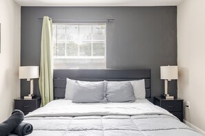 Ein modernes Schlafzimmer mit einem großen Bett, zwei Nachttischen mit Lampen, einem Fenster mit Jalousien und einer grauen Wand.