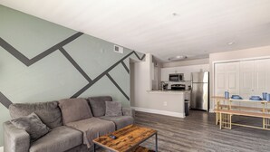 Ein modernes Wohnzimmer mit einem grauen Sofa, einem Holztisch und einer Küche im Hintergrund.