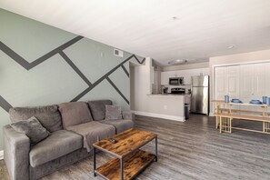 A modern living room with a grey sofa, a wooden coffee table, and a kitchen in the background. - Green King 2Bed 2Bath| walk to NRG| 1mi Med Center (Houston)
