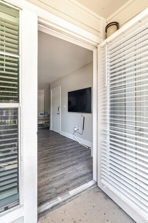 A hallway with a door leading to a room, a mounted flat-screen TV, and a window with blinds. - Green King 2Bed 2Bath| walk to NRG| 1mi Med Center (Houston)