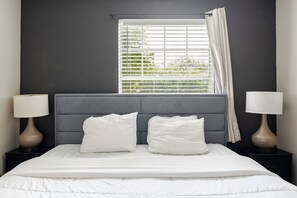 Un dormitorio con cama, dos lámparas de noche, una ventana con persianas y cabecero gris.