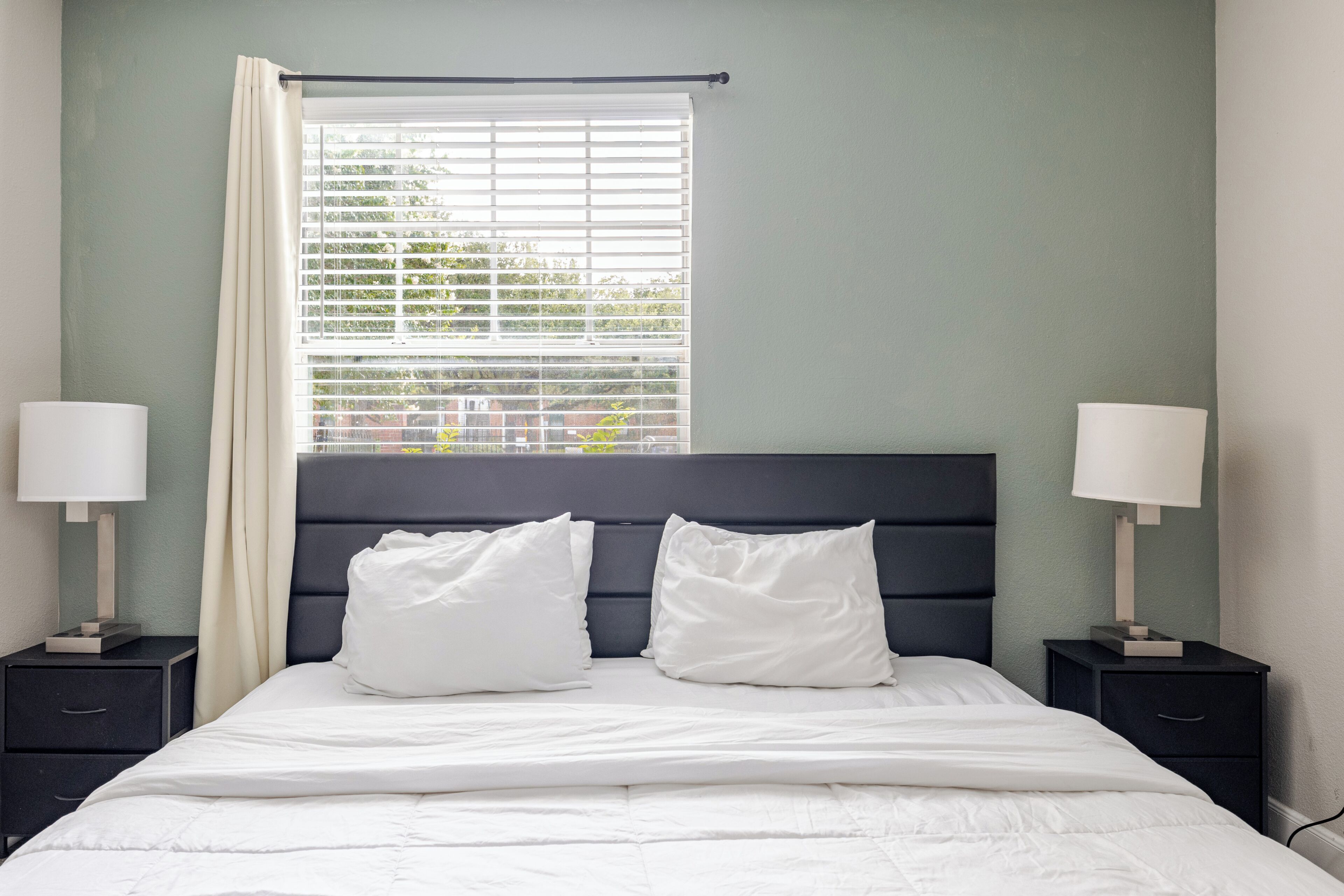 A neatly made bed with white bedding, two bedside tables with lamps, a window with blinds, and a green wall.