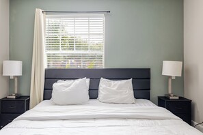 Un lit bien fait, avec une literie blanche, deux tables de chevet avec des lampes, une fenêtre avec des stores et un mur vert.