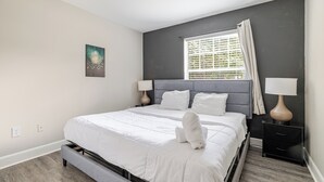 Ein modernes Schlafzimmer mit einem großen Bett, zwei Nachttischen mit Lampen, einem Fenster mit Jalousien und einem einzelnen Kunstwerk an der Wand.