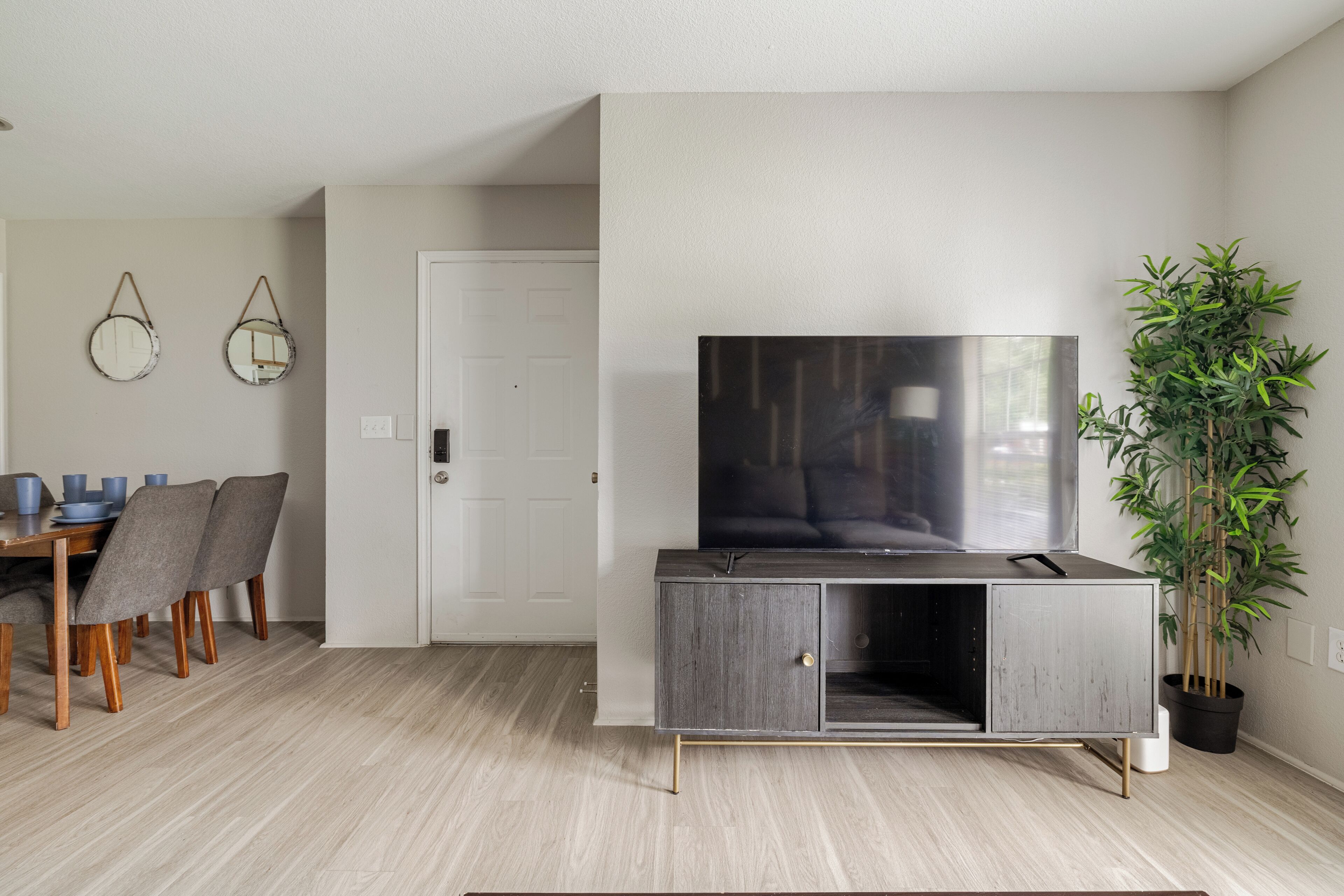 A modern living room with a grey cabinet, a flat-screen TV, a dining table with chairs, and a potted plant.