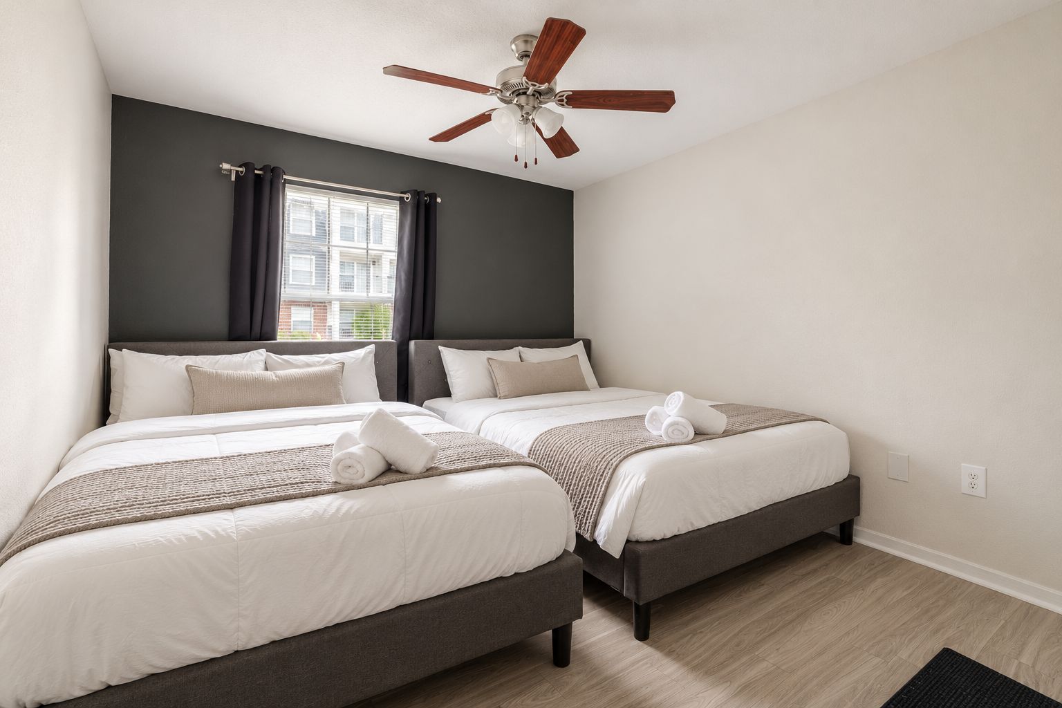 A hotel room with two beds, a ceiling fan, a window with curtains, and a view of a building outside.