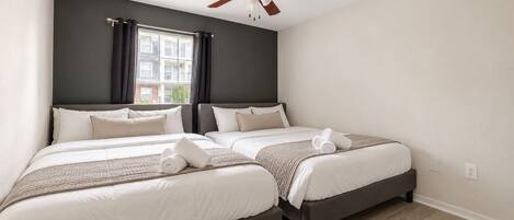 A hotel room with two beds, a ceiling fan, a window with curtains, and a view of a building outside.