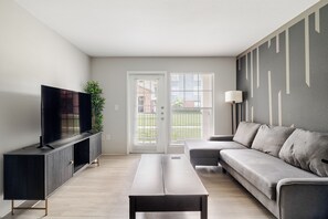A modern living room with a flat-screen TV on a black cabinet, a grey sofa, a wooden coffee table, and a large window with blinds. - Tidy 1BR with 2 Beds /Walk to NRG /By Med Center (Houston)