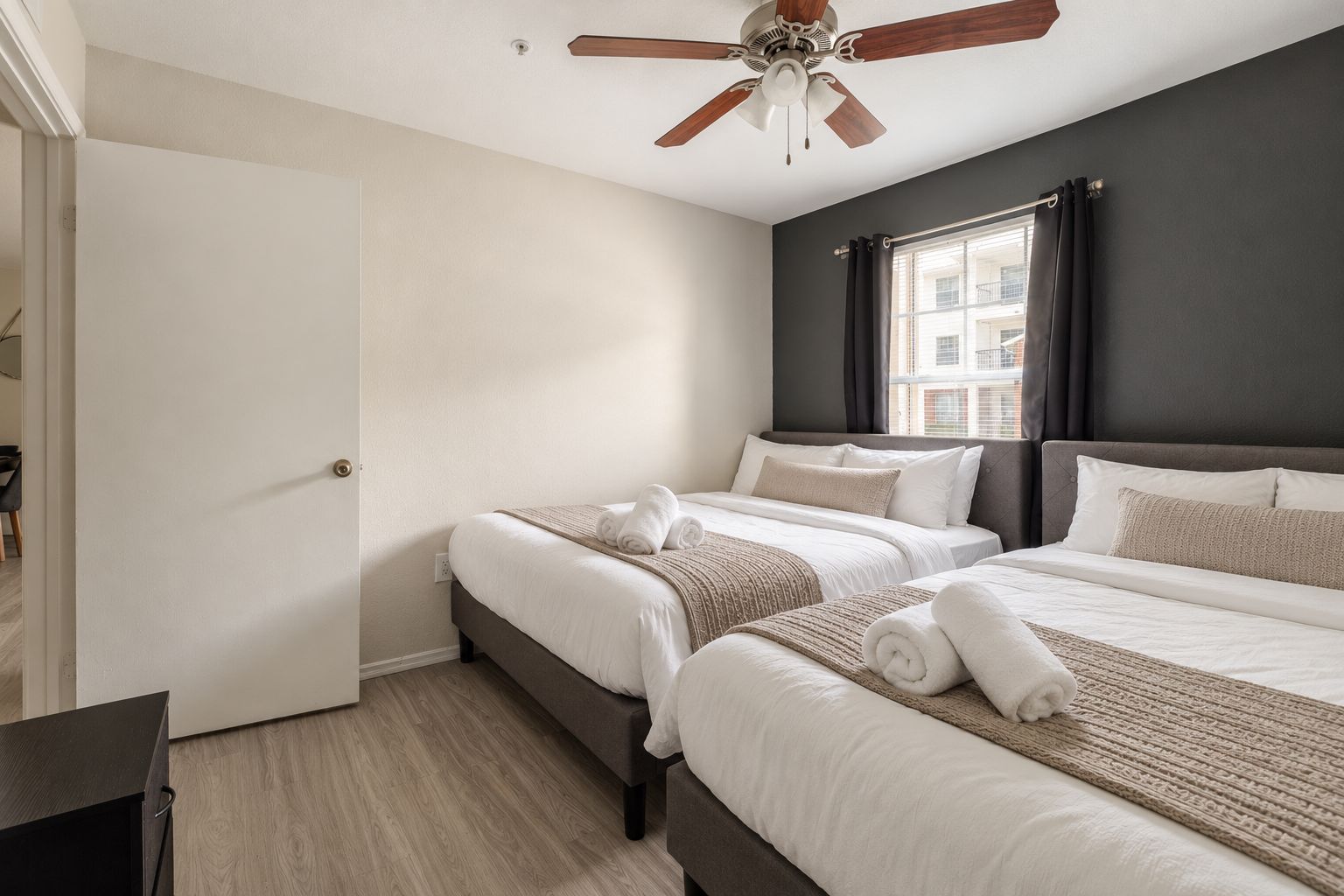 A hotel room with two beds, a ceiling fan, a window with curtains, and a view of buildings outside.