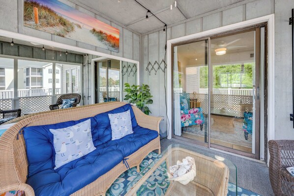A wicker sofa with blue cushions, a glass coffee table, and a wicker basket on a porch with a view of a deck and greenery.
