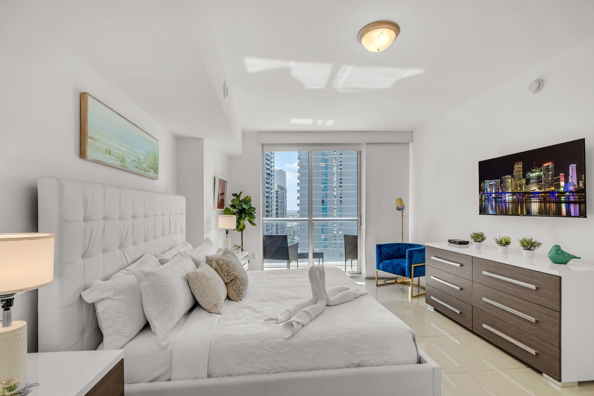 Une chambre moderne avec un grand lit, une vue sur la ville, un bureau avec des tiroirs et une chaise bleue.