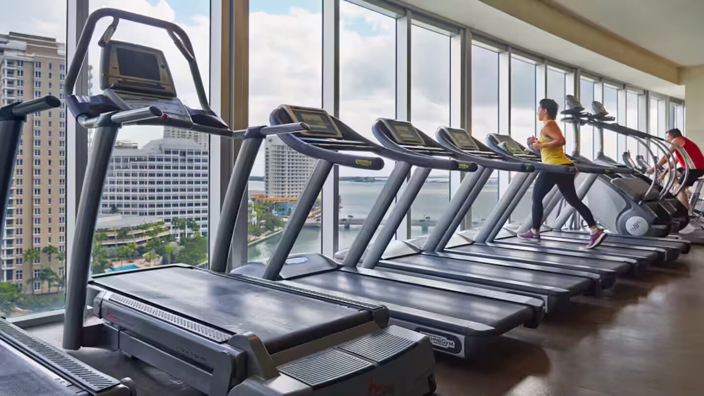 Ein Fitnessraum mit mehreren in einer Reihe angeordneten Laufbändern, großen Fenstern und Blick auf eine Stadtlandschaft.
